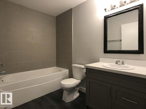 Bathroom featuring a tiled shower surround, white bathtub, wood-finish flooring, gray vanity with white countertop, and framed mirror - 13309 128 Street Nw, Edmonton, AB 