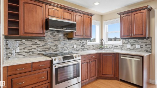 Kitchen featuring wood-finish cabinetry, light-toned countertops, a full tile backsplash, stainless steel appliances, and wood-finish flooring - 6116 53 Avenue, Beaumont, AB - Indoor Photo Showing Kitchen With Stainless Steel Kitchen With Double Sink With Upgraded Kitchen