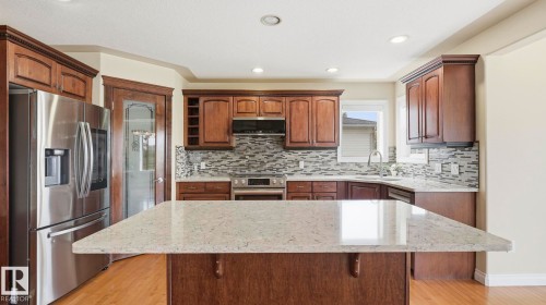 Kitchen featuring a central island with a light-toned countertop, wood-finish cabinetry, stainless steel appliances, a tiled backsplash, and recessed lighting - 6116 53 Avenue, Beaumont, AB - Indoor Photo Showing Kitchen With Upgraded Kitchen