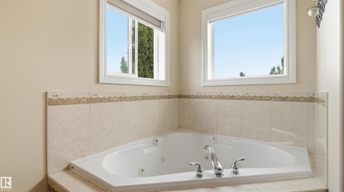 Corner built-in jetted tub with chrome fixtures, surrounded by neutral-toned tiled walls with a decorative border, and illuminated by two rectangular windows with white frames - 6116 53 Avenue, Beaumont, AB - Indoor Photo Showing Bathroom