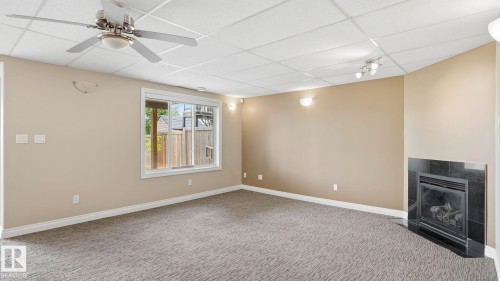 Recessed ceiling featuring a ceiling fan, carpeted flooring, a black tiled fireplace, and a window providing exterior visibility - 6116 53 Avenue, Beaumont, AB - Indoor Photo Showing Other Room With Fireplace