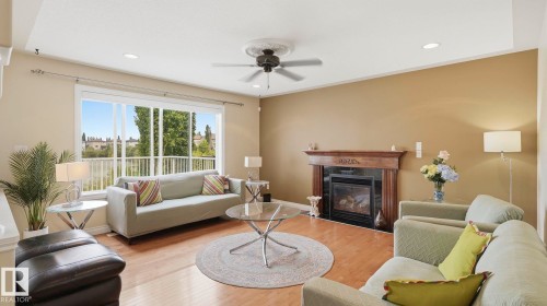 Living area featuring a large window with white framing, a wood-finish floor, a decorative fireplace with a wood mantle, a ceiling fan, and recessed lighting - 6116 53 Avenue, Beaumont, AB - Indoor Photo Showing Living Room With Fireplace