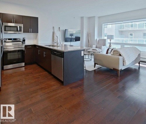 Open-concept living area featuring rich wood-finish flooring, a kitchen with dark cabinetry, stainless steel appliances, and a light-colored countertop - #605 10152 104 St Nw, Edmonton, AB 