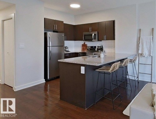Modern kitchen featuring dark wood cabinetry, stainless steel appliances, a center island with a light-toned countertop, and wood-finish flooring - #605 10152 104 St Nw, Edmonton, AB 