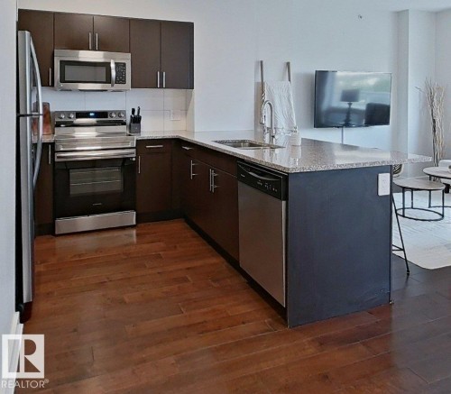 Kitchen featuring wood-finish flooring, stainless steel appliances, dark wood-finish cabinetry, and a light-toned countertop - #605 10152 104 St Nw, Edmonton, AB 