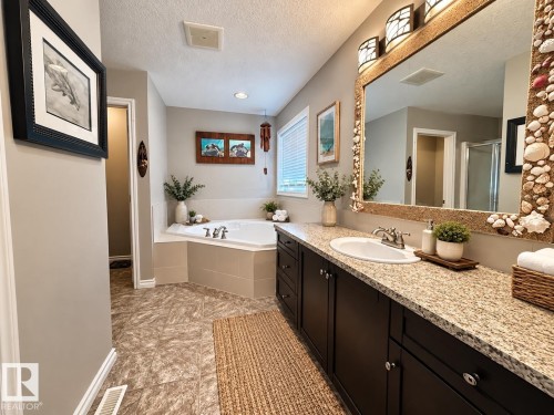 Spacious bathroom featuring a built-in corner soaking tub, dual-sink vanity with a light-toned countertop, dark wood-finish cabinetry, and patterned floor tiling - 16893 58A Street Nw, Edmonton, AB 