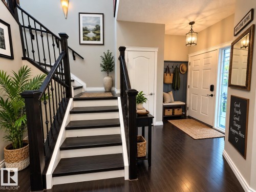 Two-story foyer featuring dark wood-finish flooring, a grand staircase with wrought iron spindles, a front entry door with a sidelight, and an integrated built-in bench with hooks - 16893 58A Street Nw, Edmonton, AB 