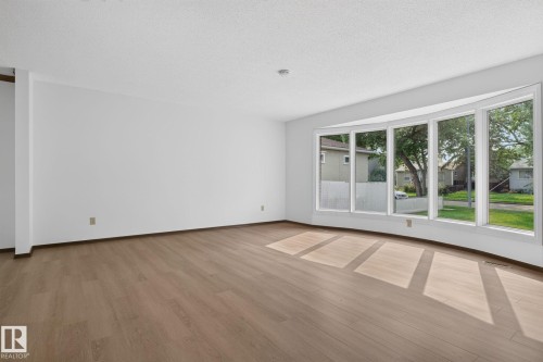 Expansive living space featuring a five-panel bay window, light wood-finish flooring, white walls, and a textured white ceiling - 12121 42 Street Nw, Edmonton, AB - Indoor Photo Showing Other Room
