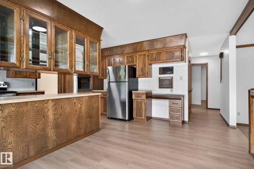 Kitchen featuring wood-finish cabinetry, integrated glass-front display cabinets, light-toned countertops, tile backsplash, and wood-finish flooring - 12121 42 Street Nw, Edmonton, AB - Indoor Photo Showing Kitchen With Stainless Steel Kitchen