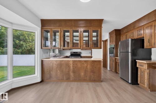 Kitchen featuring extensive wood cabinetry, a bay window overlooking a green space, stainless steel refrigerator, built-in wall oven, and light wood-finish flooring - 12121 42 Street Nw, Edmonton, AB - Indoor Photo Showing Kitchen
