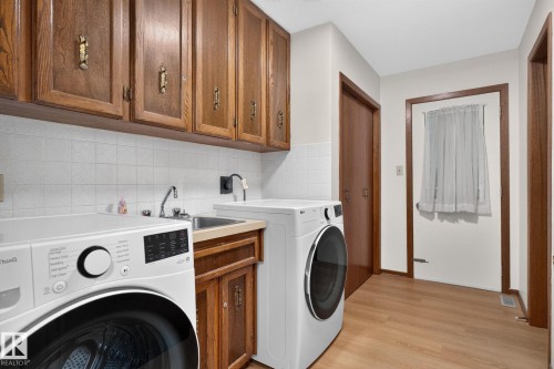 Dedicated laundry room featuring wood-finish flooring, wood cabinetry with decorative hardware, a utility sink, and white tile backsplash - 12121 42 Street Nw, Edmonton, AB - Indoor Photo Showing Laundry Room