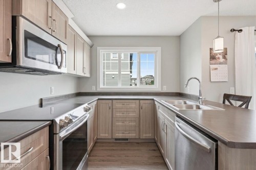 Modern kitchen featuring custom wood-finish cabinetry, stainless steel appliances including a built-in microwave and dishwasher, gray quartz countertops, a chrome single-handle faucet, and wood-finish flooring throughout - #12 1391 Starling Dr Nw, Edmonton, AB 