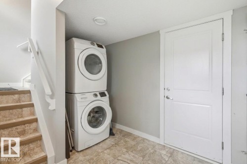 Integrated laundry area featuring a stacked washer and dryer, adjacent to a staircase with wood-finish treads and a white handrail - #12 1391 Starling Dr Nw, Edmonton, AB 