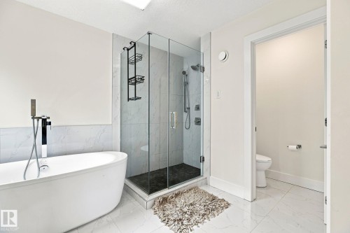 Freestanding soaking tub with floor-mounted faucet, glass-enclosed shower with dark tile base and wall-mounted shower system, and large format tile flooring - 4530 Saly Place Sw, Edmonton, AB 