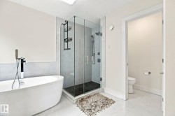Freestanding soaking tub with floor-mounted faucet, glass-enclosed shower with dark tile base and wall-mounted shower system, and large format tile flooring - 
