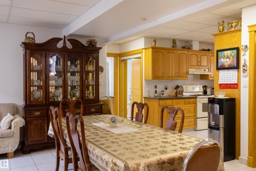Open-concept dining area with tile flooring and a dark wood china cabinet, adjacent to a kitchen featuring wood-finish cabinetry, white appliances, and a tiled backsplash - 6407 163 Avenue Nw, Edmonton, AB 