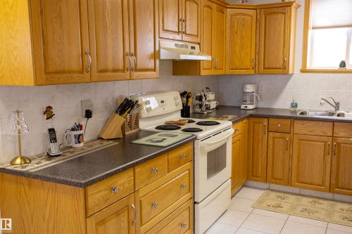 Kitchen featuring wood-finish cabinetry with silver-tone hardware, dark countertops, a white tile backsplash, and a built-in white electric range - 6407 163 Avenue Nw, Edmonton, AB 