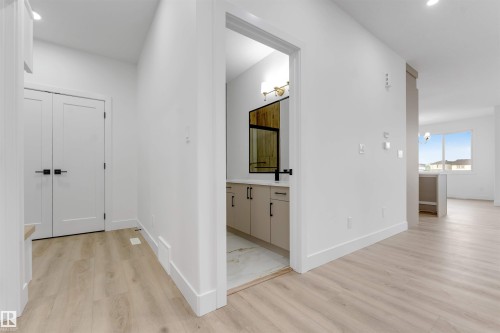 Bright interior featuring wood-finish flooring, white walls, and a visible powder room with a vanity, rectangular mirror, and wall-mounted light fixture - 4234 63 Street, Beaumont, AB - Indoor Photo Showing Other Room