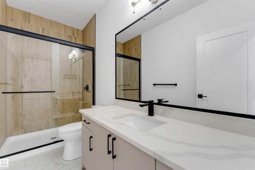 Bathroom featuring a frameless glass shower enclosure with matte black hardware, floor-to-ceiling wood-look tile, a single vanity with stone-finish countertop, under-mount sink, and matte black faucet, and white marble-look floor tiling - 4234 63 Street, Beaumont, AB - Indoor Photo Showing Bathroom