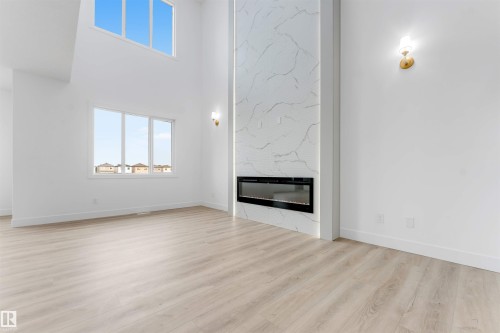Two-story great room featuring a tall marble-look fireplace with an integrated linear electric insert, light wood-finish flooring, and multiple large windows for abundant illumination - 4234 63 Street, Beaumont, AB - Indoor With Fireplace