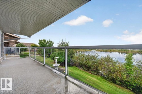 Expansive outdoor deck with a textured surface, glass railing, and a covered ceiling, overlooking a body of water with lush landscaping - #7 10500 103 Av, Morinville, AB 