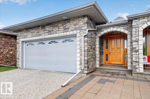 Stone facade with arched entryway, natural wood front door, and a paneled garage door with decorative windows - #7 10500 103 Av, Morinville, AB 
