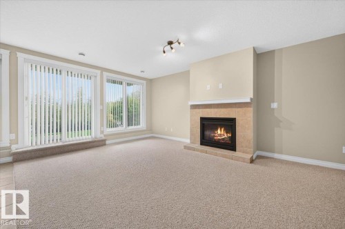 Living area featuring a gas fireplace with a tiled surround, neutral carpeting, and multiple windows with vertical blinds - #7 10500 103 Av, Morinville, AB 