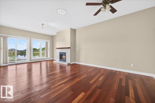 High-gloss wood-finish flooring, a fireplace with a tile surround and wood mantel, multiple windows providing natural light, a ceiling fan, and recessed lighting - #7 10500 103 Av, Morinville, AB 