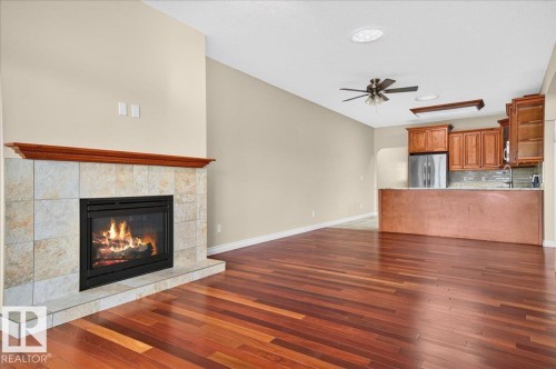 Hardwood flooring throughout, tile-surround fireplace with wood mantel, ceiling fan, wood cabinetry, and stainless steel refrigerator - #7 10500 103 Av, Morinville, AB 