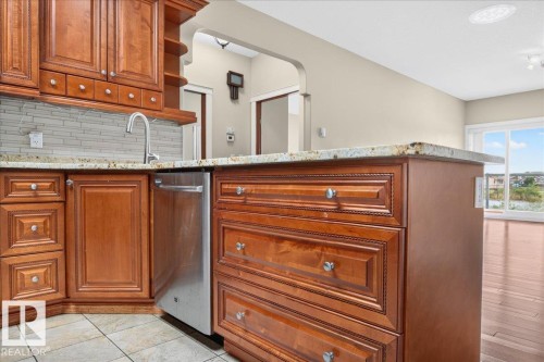 Warm wood cabinetry, integrated stainless steel dishwasher, light-toned tile backsplash, and stone countertops - #7 10500 103 Av, Morinville, AB 