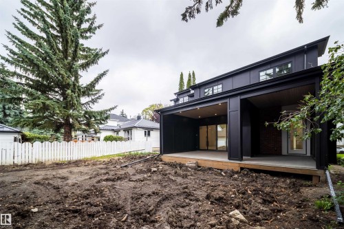 Modern black exterior with a sloped roofline, featuring two covered patios with recessed lighting, a white picket fence, and mature evergreen trees - 10414 134 Street Nw, Edmonton, AB 