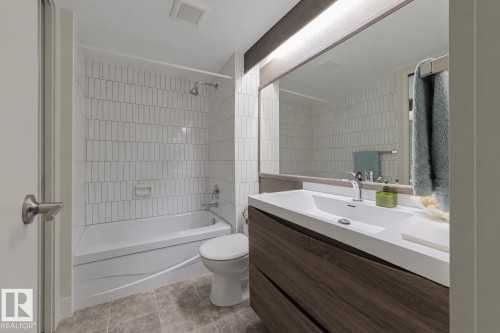 Bathroom featuring a white bathtub with subway tile surround, a toilet, a wood-finish vanity with an integrated sink, and a large wall-mounted mirror with overhead lighting - #2005 10149 Saskatchewan Dr Nw, Edmonton, AB 