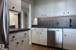 White louvered cabinetry with black hardware, stainless steel dishwasher, dark countertop, and dark tiled backsplash with under-cabinet lighting - 