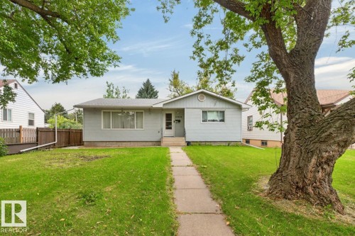 Single-story residence featuring light-colored siding, a mature front yard tree, a concrete walkway, and a partial wood fence - 12424 77 Street Nw, Edmonton, AB 