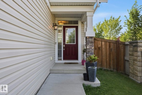 Welcoming entry featuring a rich burgundy door with clear pane window, exterior sconce lighting, stone-clad pillar, and an expansive concrete walkway - 3204 Abbott Crescent Sw, Edmonton, AB - Outdoor With Exterior