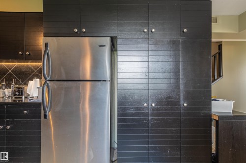 Integrated stainless steel refrigerator, dark wood-finish cabinetry, black tiled backsplash, and recessed lighting - #1202 10010 119 St Nw, Edmonton, AB 