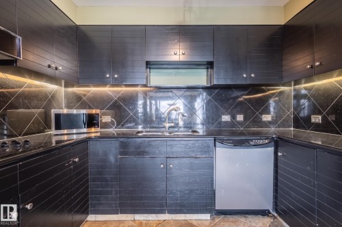 U-shaped kitchen featuring black cabinetry, dark countertops, a tile backsplash with geometric pattern, and a stainless steel dishwasher - #1202 10010 119 St Nw, Edmonton, AB 