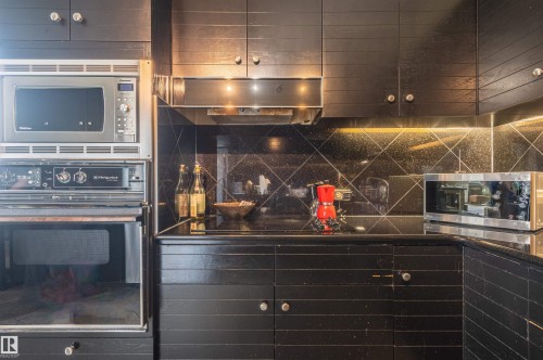 Black cabinetry with silver-tone hardware, built-in stainless steel microwave and oven, black countertop with a speckled finish, black tile backsplash with a diagonal pattern, and an under-cabinet range hood - #1202 10010 119 St Nw, Edmonton, AB 