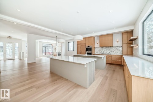 Open-concept kitchen with light wood-finish flooring, two kitchen islands featuring light countertops, wood-finish cabinetry, built-in stainless steel appliances, and a marble-patterned backsplash - 10514 134 Street Nw, Edmonton, AB 
