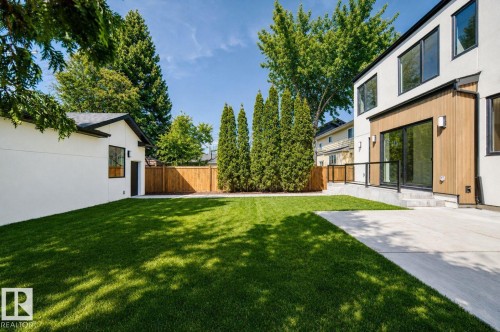 Expansive manicured lawn with a concrete patio, featuring a modern exterior with wood paneling and large windows, surrounded by mature trees and a privacy fence - 10514 134 Street Nw, Edmonton, AB 