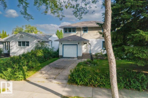 Two-story residence featuring a ground-level garage, front entrance with stairs, and a surrounding lawn with mature trees and shrubbery - 14608 93 Avenue Nw, Edmonton, AB 