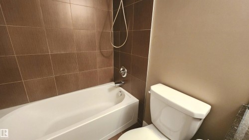 White bathtub with chrome fixtures, complemented by brown tiled shower surround and neutral painted walls - #101 11803 22 Av Sw, Edmonton, AB 