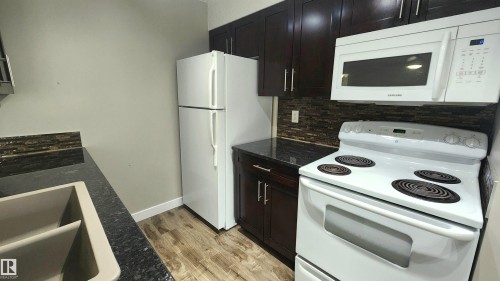 Dark wood-finish cabinetry, white appliances, dark countertops, tile backsplash, and wood-finish flooring - #101 11803 22 Av Sw, Edmonton, AB 