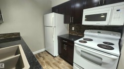 Dark wood-finish cabinetry, white appliances, dark countertops, tile backsplash, and wood-finish flooring - 