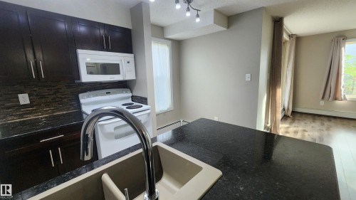 Kitchen featuring dark wood-finish cabinetry, white built-in microwave, white electric range, dark stone-finish countertops, and a dual-basin sink with a chrome faucet - #101 11803 22 Av Sw, Edmonton, AB 