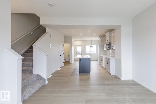 Open-concept living space featuring wood-finish flooring, a contemporary kitchen with light cabinetry, a dark kitchen island with an integrated sink, and a staircase with neutral-toned carpeting - 70 Dansereau Way, Beaumont, AB - Indoor
