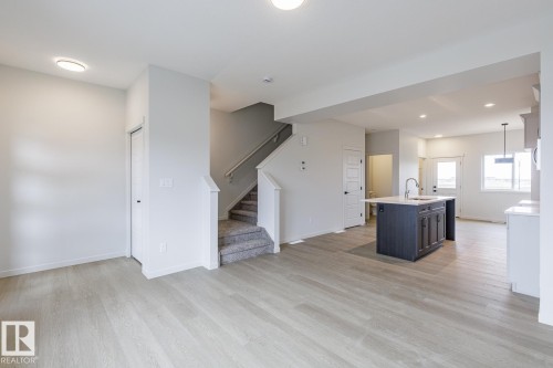 Open-concept living area featuring light wood-finish flooring, recessed ceiling lighting, a dark-toned kitchen island with a sink, white perimeter cabinetry, and a carpeted staircase with a handrail - 70 Dansereau Way, Beaumont, AB - Indoor