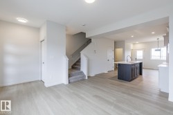 Open-concept living area featuring light wood-finish flooring, recessed ceiling lighting, a dark-toned kitchen island with a sink, white perimeter cabinetry, and a carpeted staircase with a handrail - 