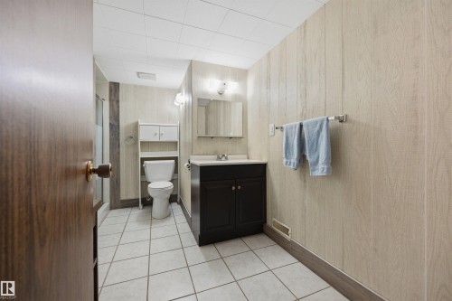 White tiled flooring, dark wood-finish vanity with white countertop, wall-mounted mirror, and wood-paneled walls - 14008 94 Street Nw, Edmonton, AB 