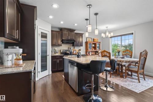 Kitchen featuring wood-finish flooring, dark wood cabinetry, light granite countertops, stainless steel appliances, and recessed lighting - 5130 Godson Close Nw, Edmonton, AB 